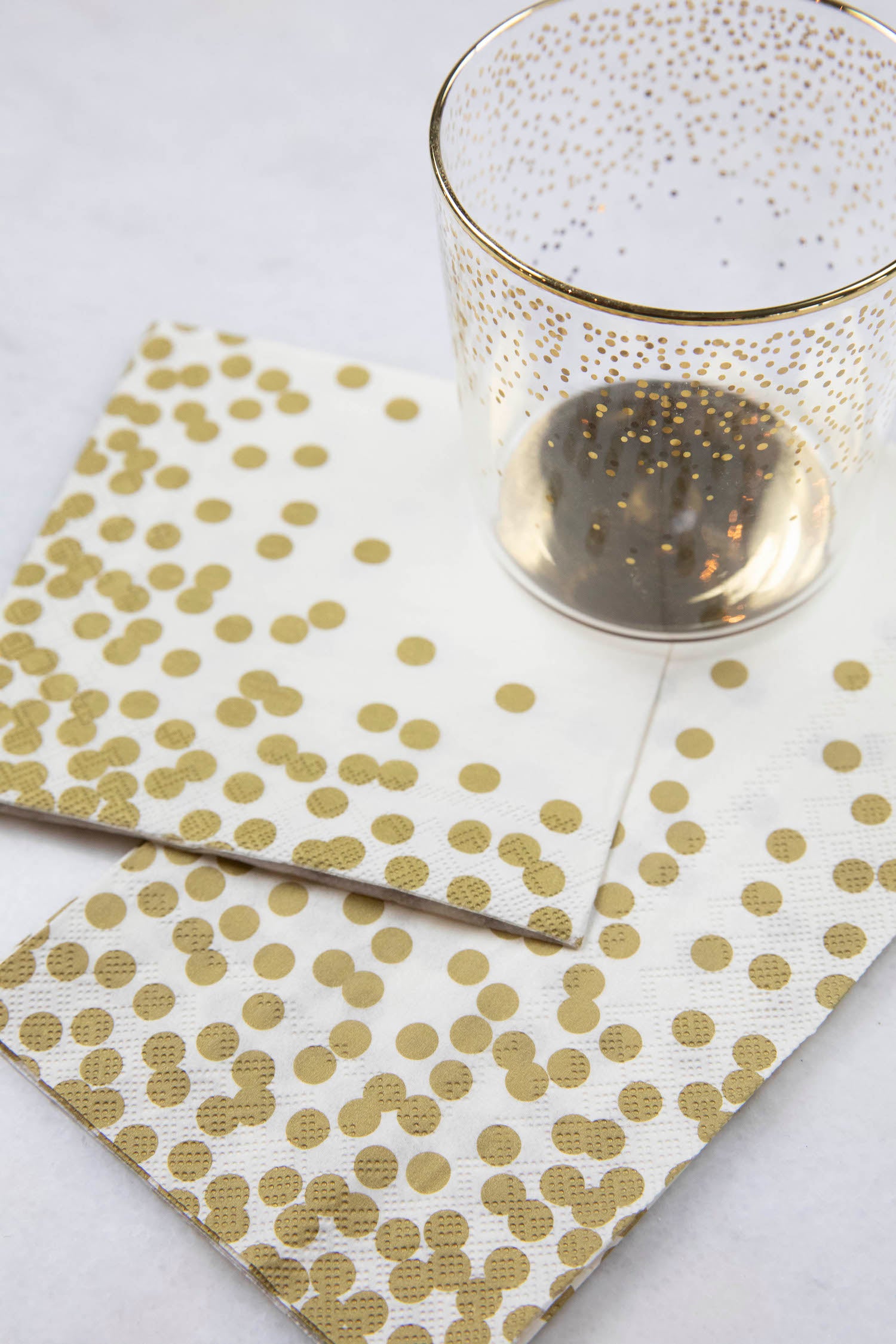Two Gold Confetti Napkins, one Cocktail and one Guest, under a gold-accented glass on a white table.