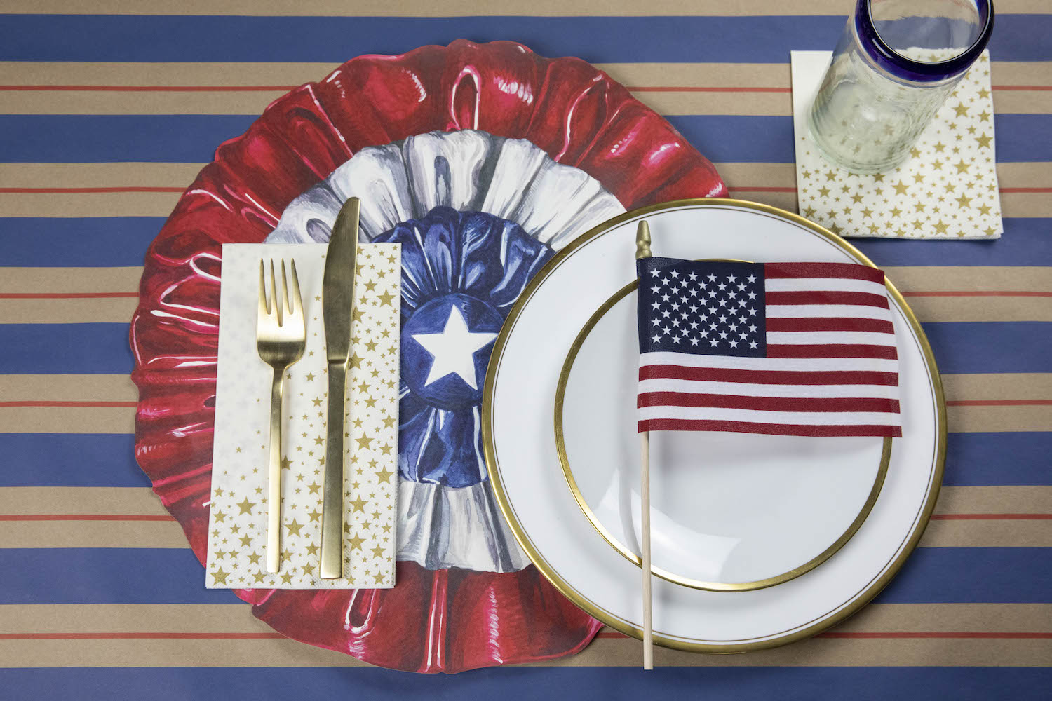 A patriotic place-setting with a Shining Star Guest Napkin with a gold fork and knife next to the plate, and a Shining Star Cocktail Napkin under the glass.