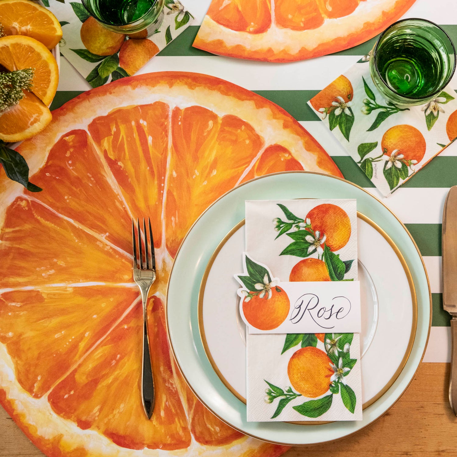 A vibrant citrus-themed place setting, featuring an Orange Orchard Guest Napkin centered on the plate, and an Orange Orchard Cocktail Napkin under the water glass.