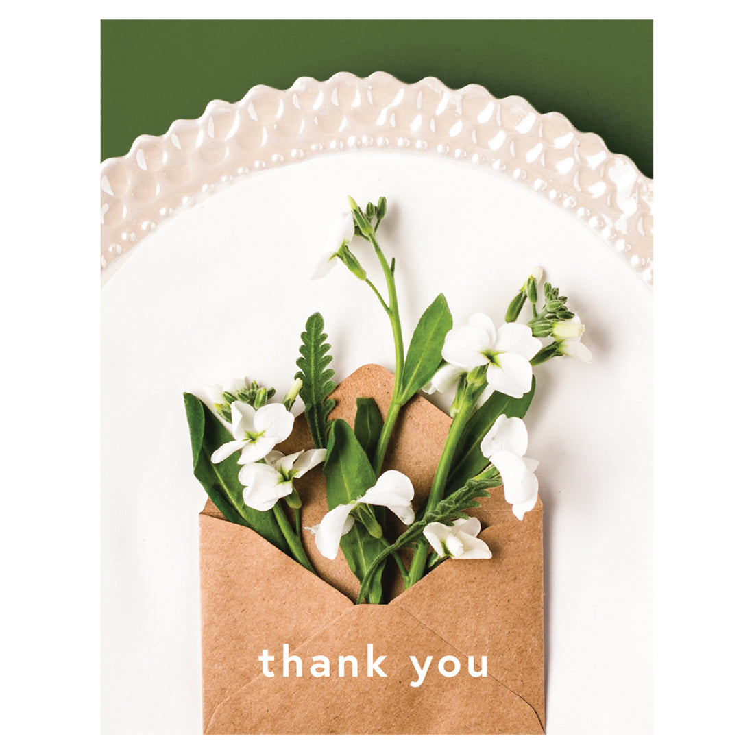 A photo of an open tan kraft paper envelope with delicate white blooms sprouting from it, resting on a white plate on a green table, with &quot;thank you&quot; printed in white on the envelope.