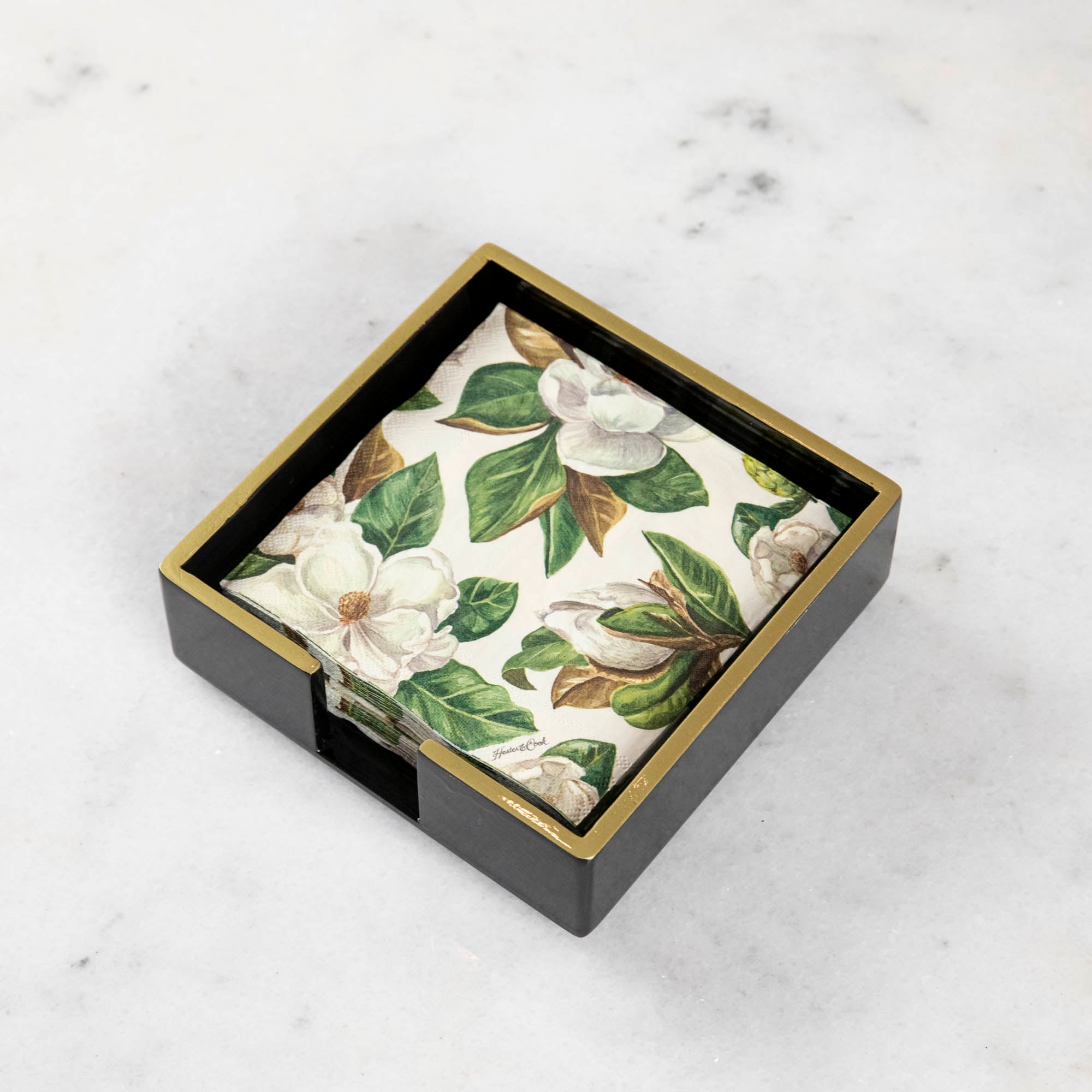 A black lacquered napkin holder with gold trim and a magnolia print napkin inside of it.