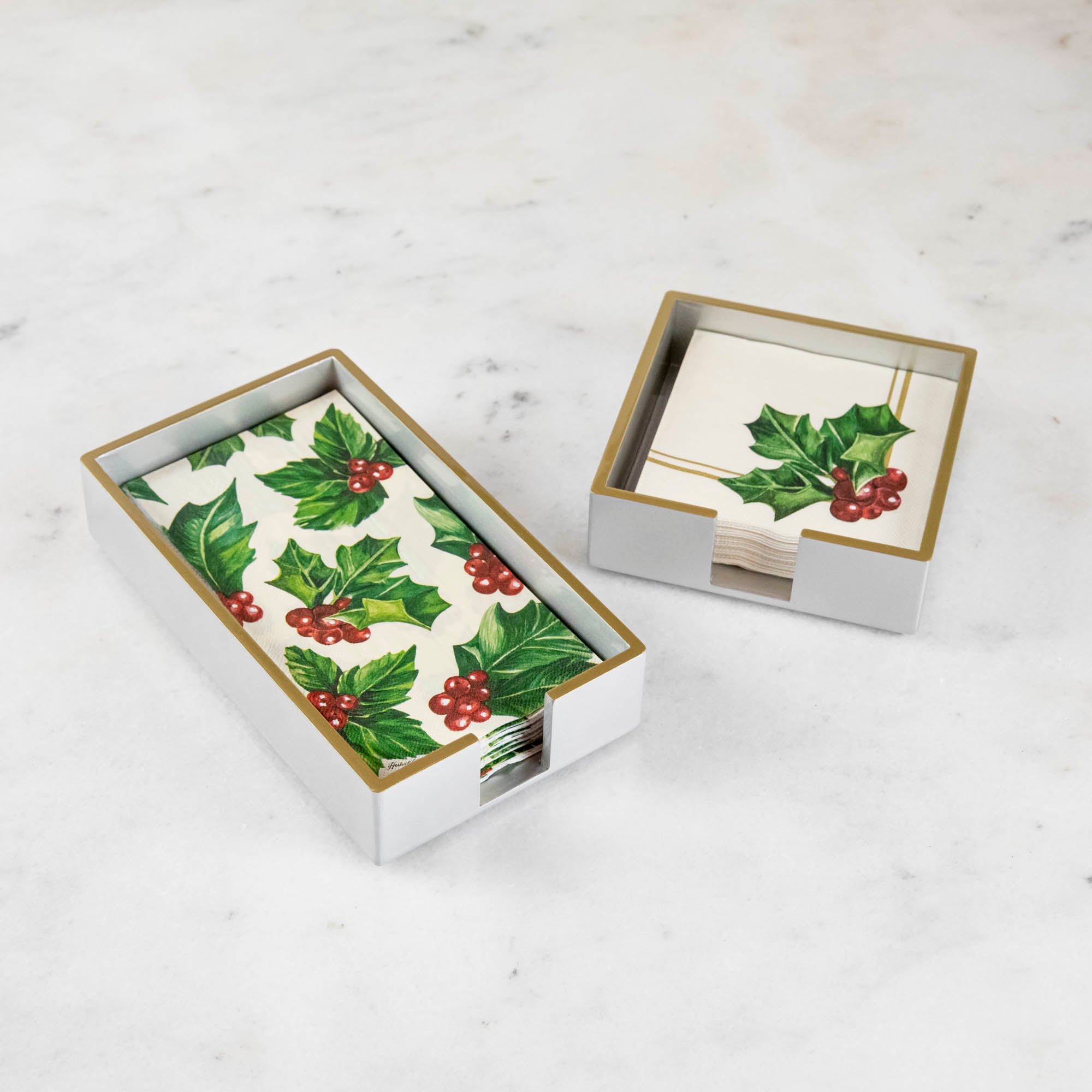 A silver colored lacquered napkin holder in guest and cocktail size with napkins in them.