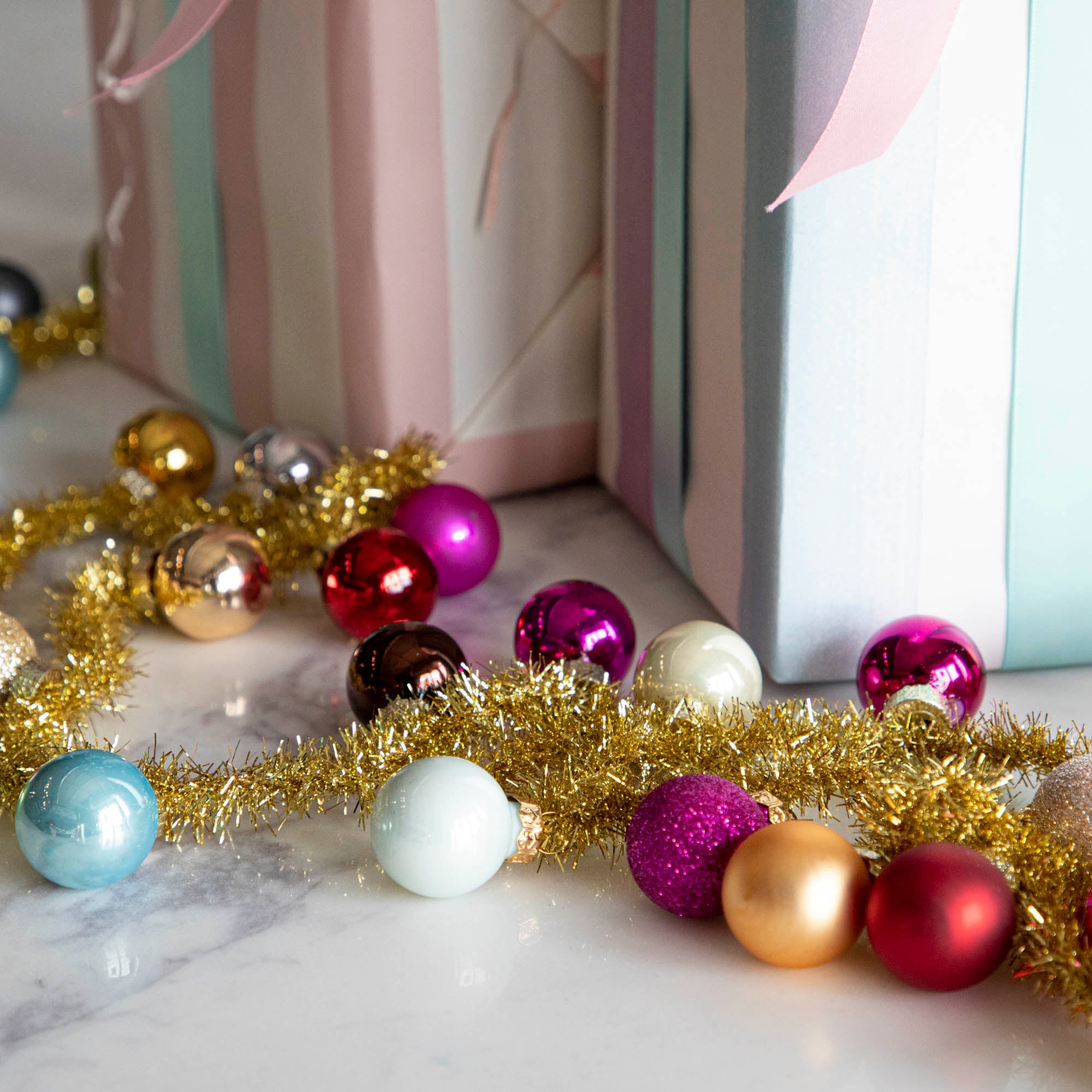 A garland made with gold tinsel string with multi colored balls attached to it strewn in front of 2 decorated gifts.