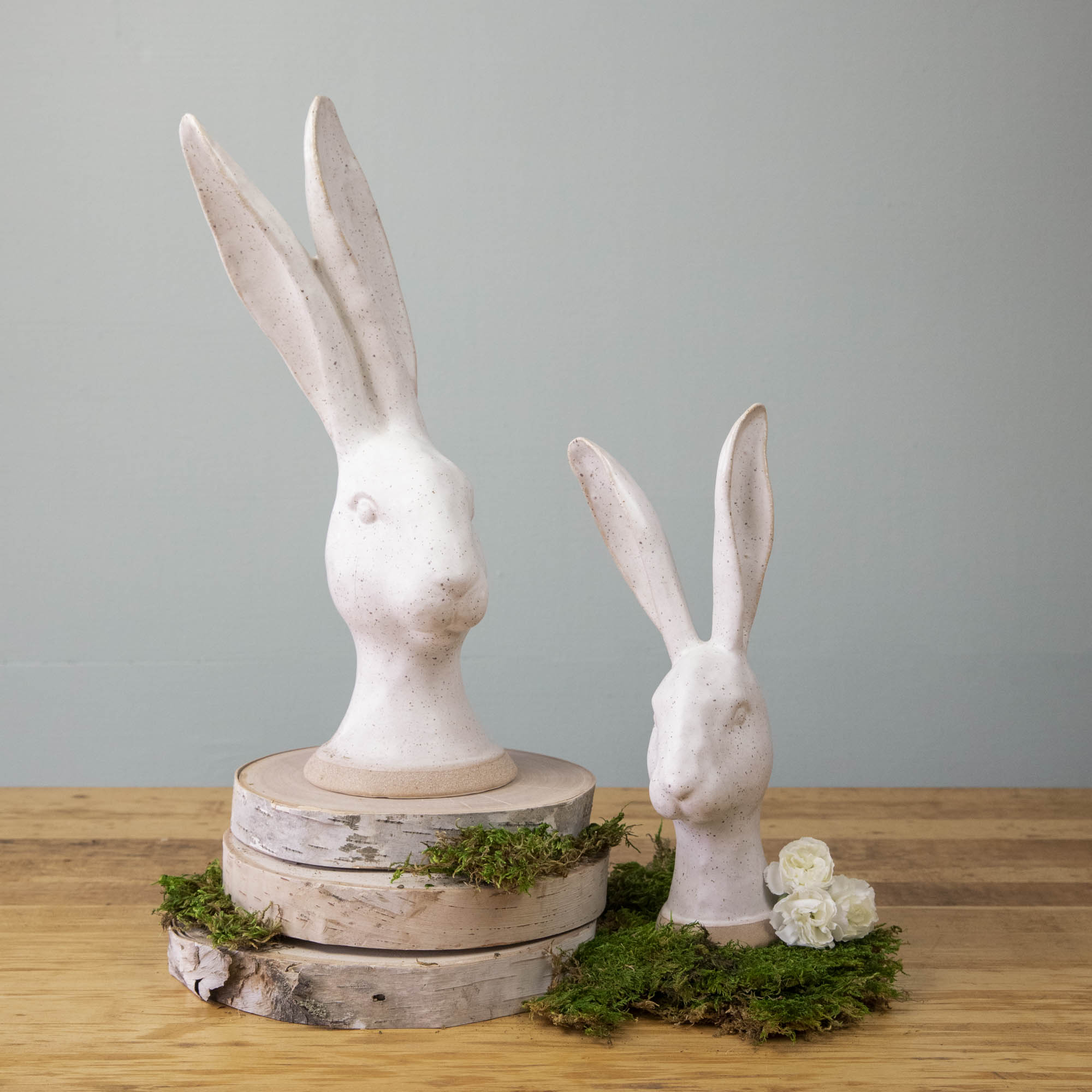 Description: Enchanting HomArt Matte White Ceramic Hares, two ceramic rabbit garden decorations, perched on top of a pile of moss.