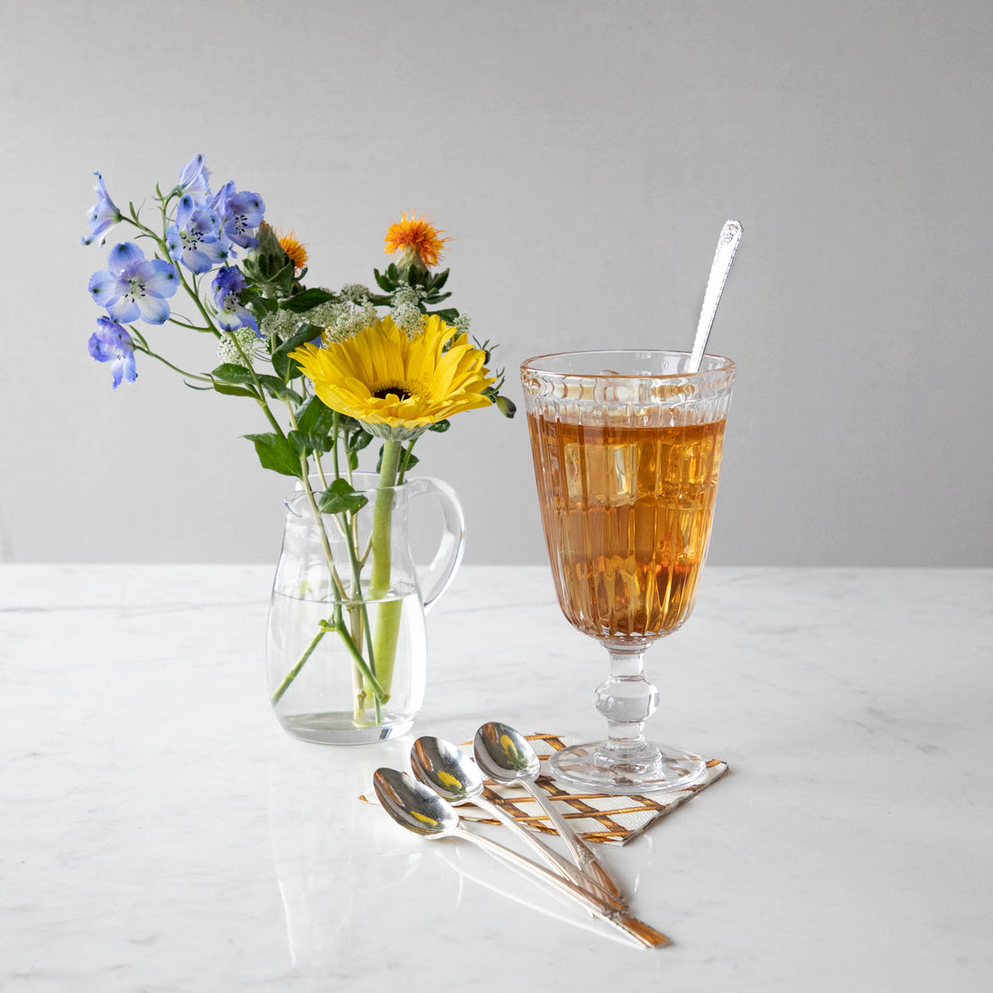 A clear glass of iced tea with a Hester &amp; Cook Vintage Silver-Plate Long Handled Tea Spoon Set of Four alongside a bouquet of colorful flowers in a clear vase, with additional vintage silver-plate teaspoons scattered nearby.