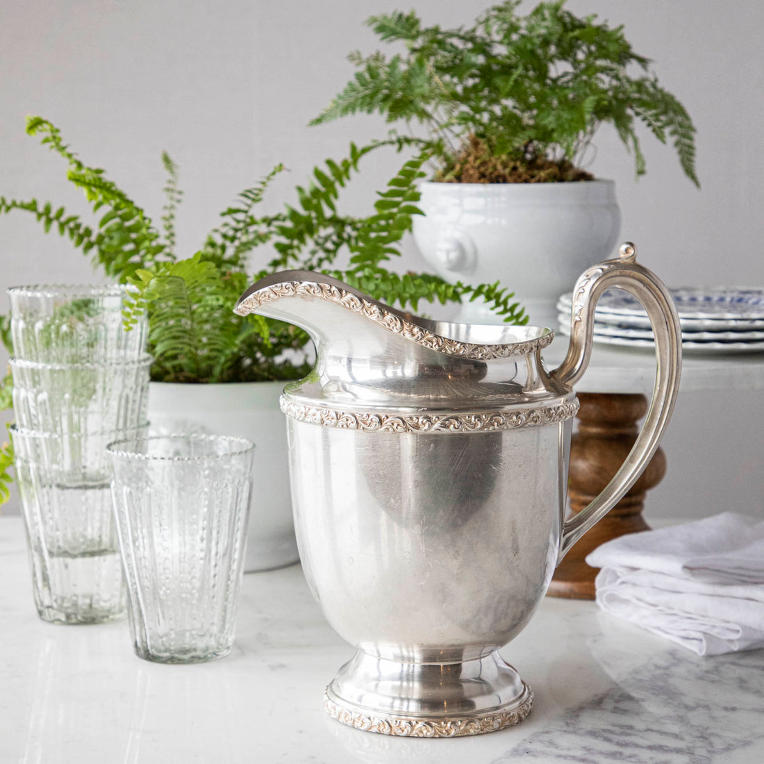 Elegant Hester &amp; Cook vintage silver-plate water pitcher on a table adorned with glasses, plates, and a potted fern in the background.