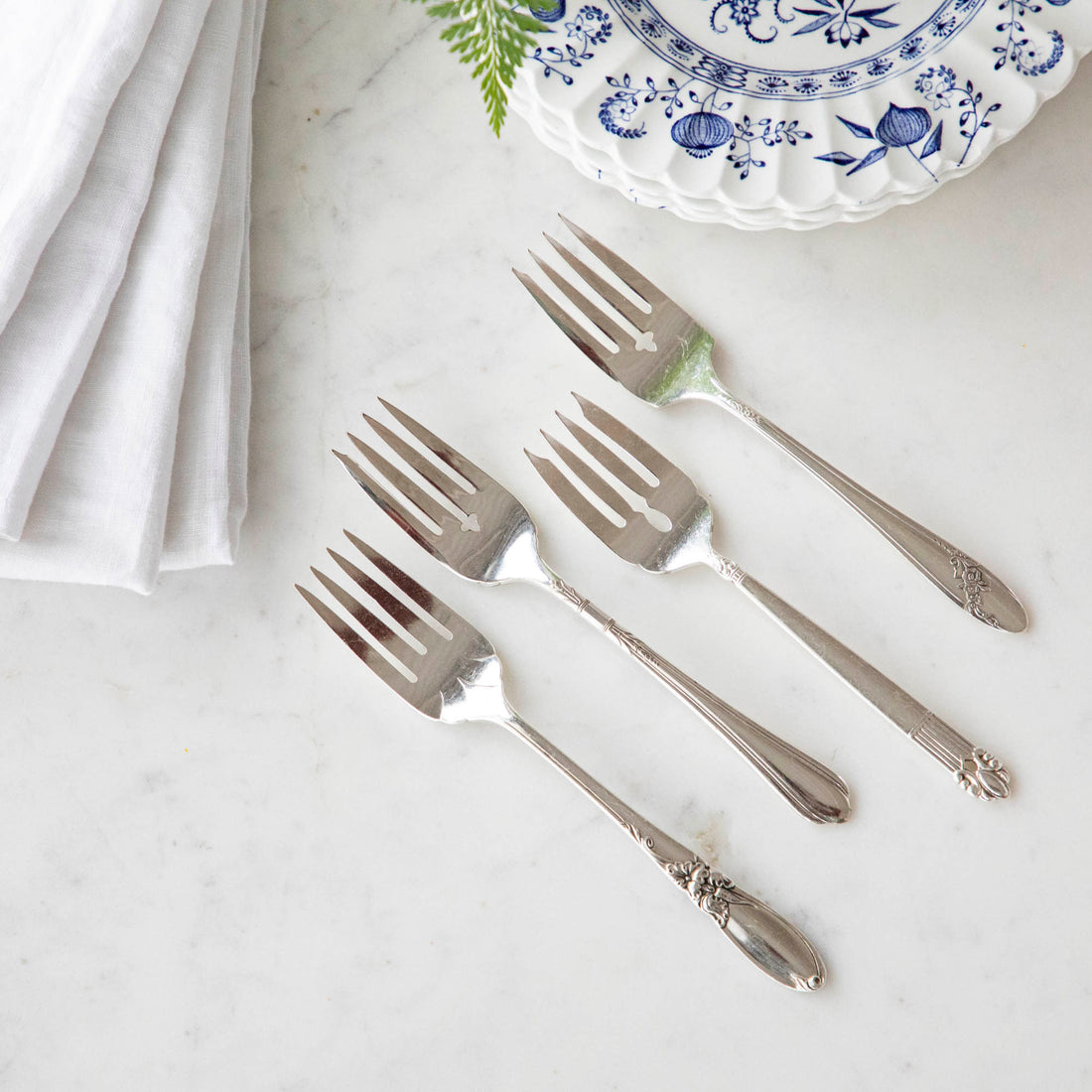 Four Hester &amp; Cook vintage silver-plate salad forks beside a blue and white patterned plate with a white napkin and a sprig of greenery.