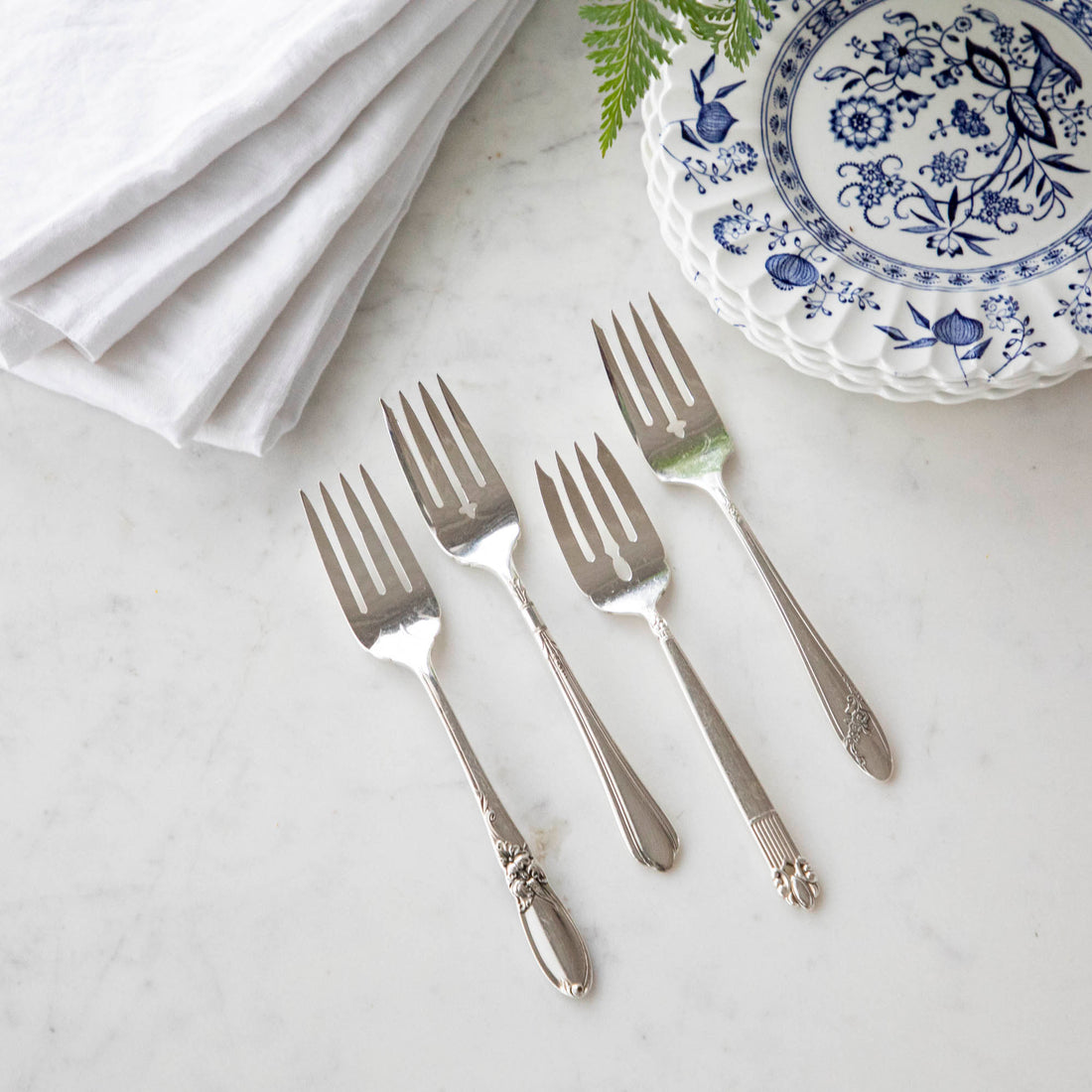 Four Hester &amp; Cook vintage silver-plate salad forks beside a blue and white patterned plate with a white napkin and a sprig of greenery.