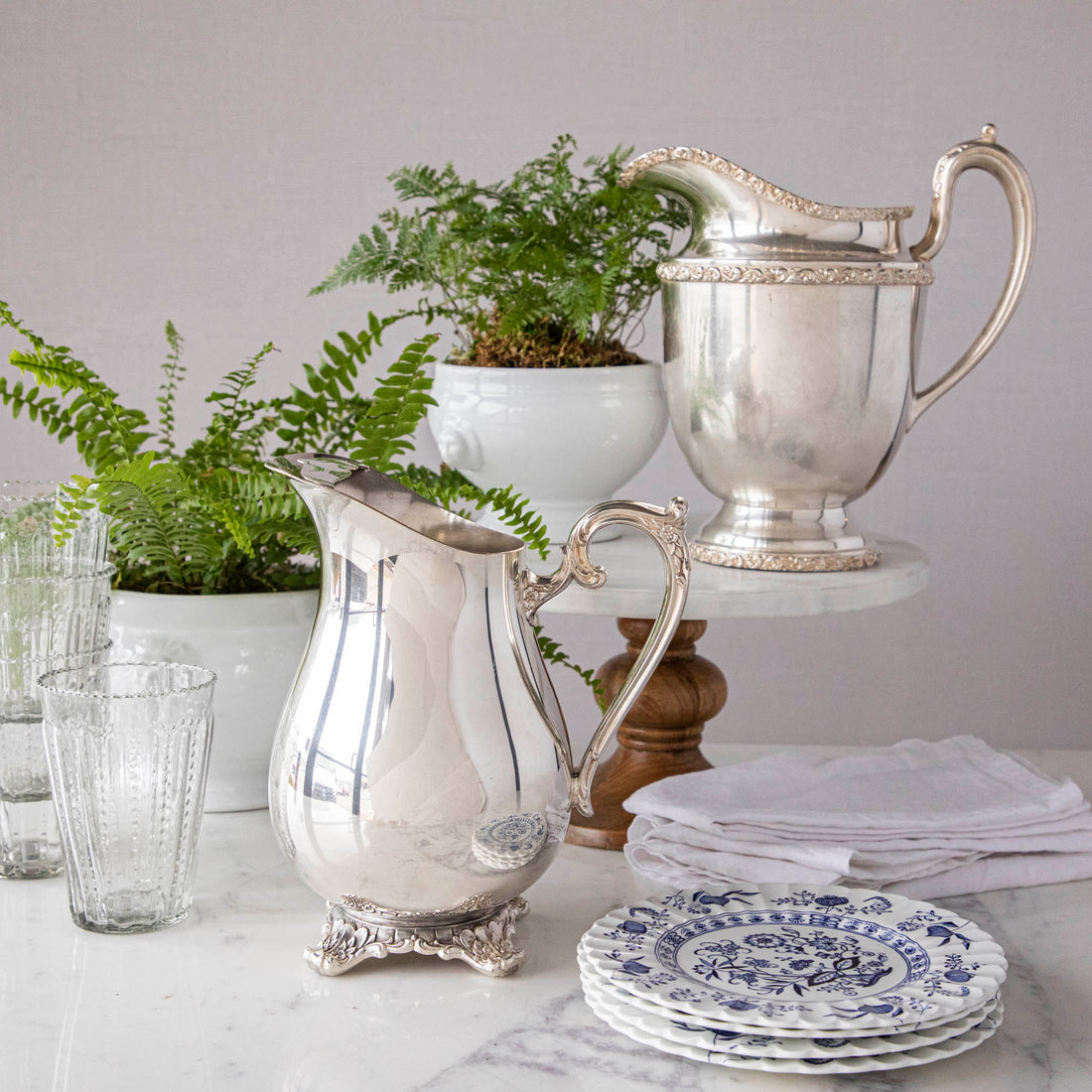 Elegant Hester &amp; Cook vintage silver-plate water pitcher on a table adorned with glasses, plates, and a potted fern in the background.