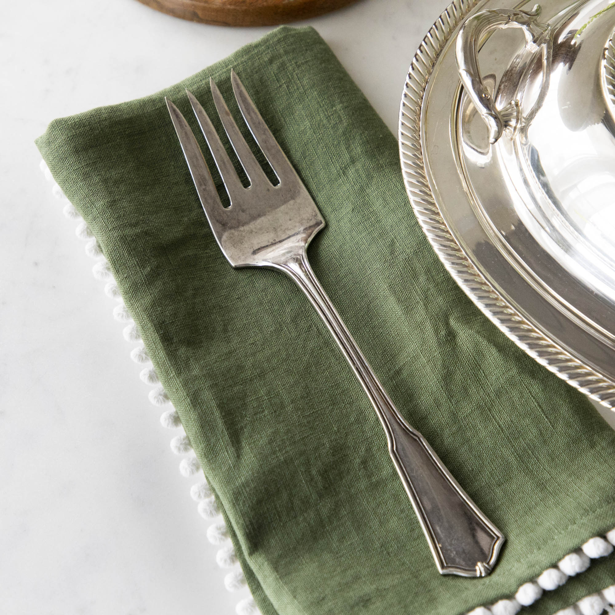A Hester & Cook vintage silver-plate meat fork resting on a folded green napkin with pom-pom trim on a marble countertop.