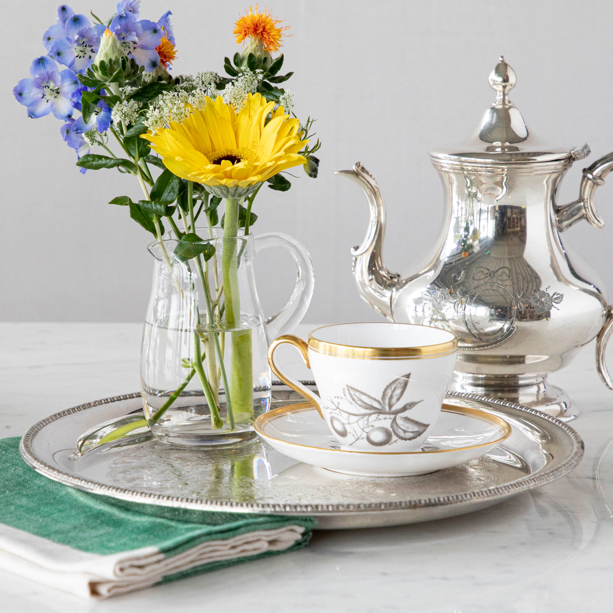 A silver-plate tea tray with a floral pattern, accompanied by a silver teapot and a cup with a floral design, presented alongside a vase with flowers.