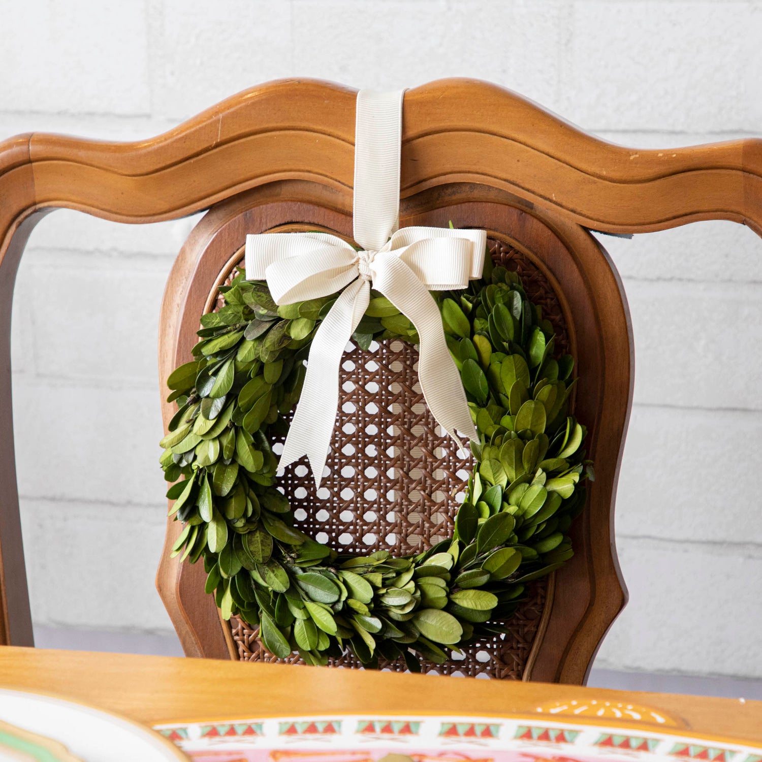 A wicker chair adorned with a Porch View Home Boxwood Wreath with Cream Grosgrain Ribbon.