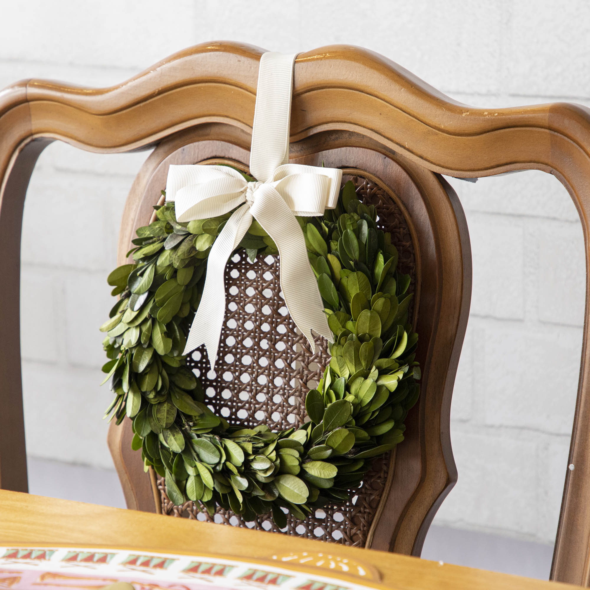 A Porch View Home Boxwood Wreath with Cream Grosgrain Ribbon hangs on the back of a wooden chair.
