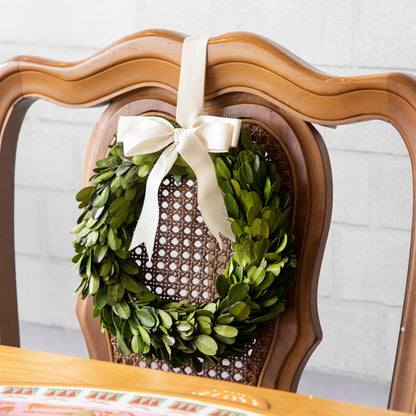 A Porch View Home Boxwood Wreath with Cream Grosgrain Ribbon hangs on the back of a wooden chair.
