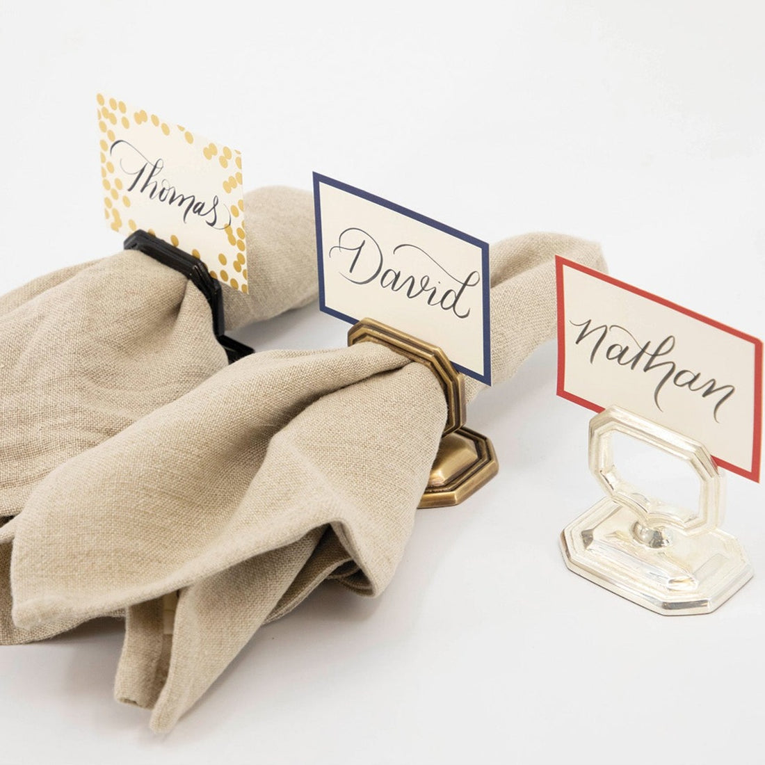 Three vintage-style Napkin Rings with Place Card Holders (Black, Brass and Silver) in a row, all three with different place cards and two holding folded cloth napkins.