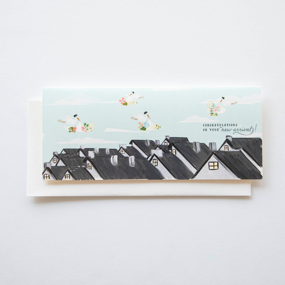 An illustration on a Rooftop Special Delivery baby arrival card from The First Snow featuring a row of houses with the text &quot;congratulations on your new arrival!&quot; above them, surrounded by images of flowers and storks in the sky.