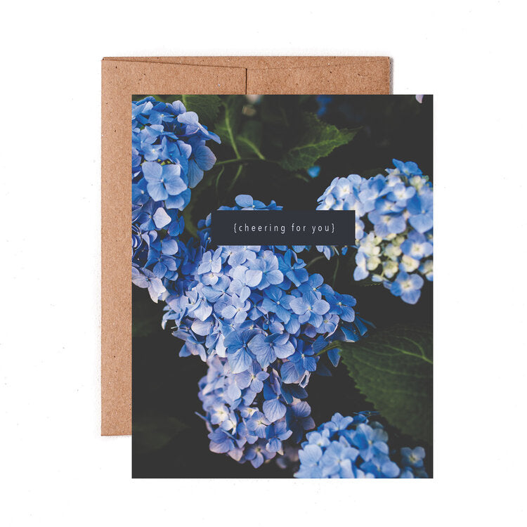 A folded Cheering For You Hydrangea Card with blue hydrangea flowers and the text &quot;{cheering for you}&quot; placed on a beige background, printed on high quality uncoated stock by The Pen + Piper.