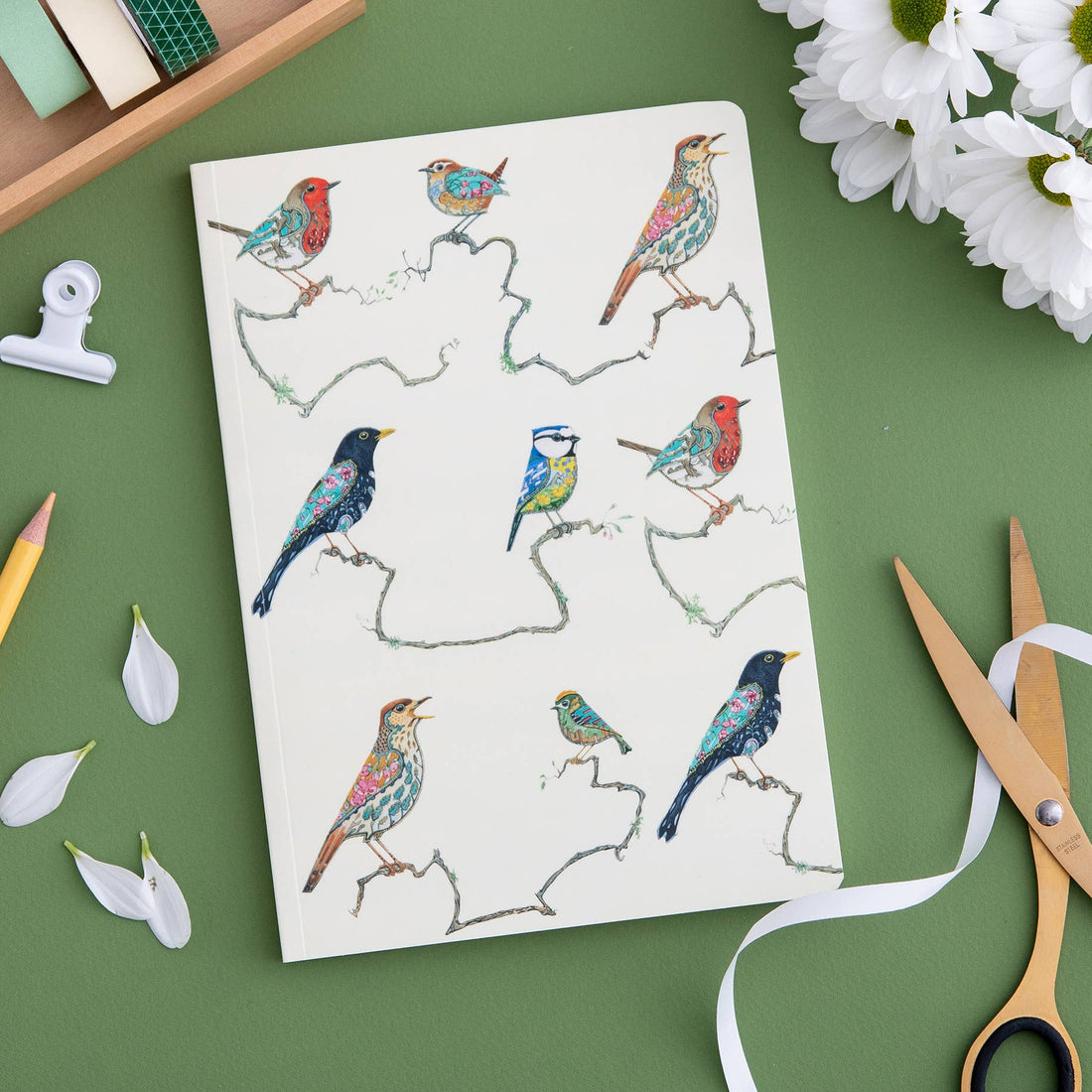 A Songbird Perfect Bound Notebook with a bird illustration on the cover surrounded by art supplies and white flowers on a green background, crafted by British papermakers on luxury cream stock, created by The DM Collection.
