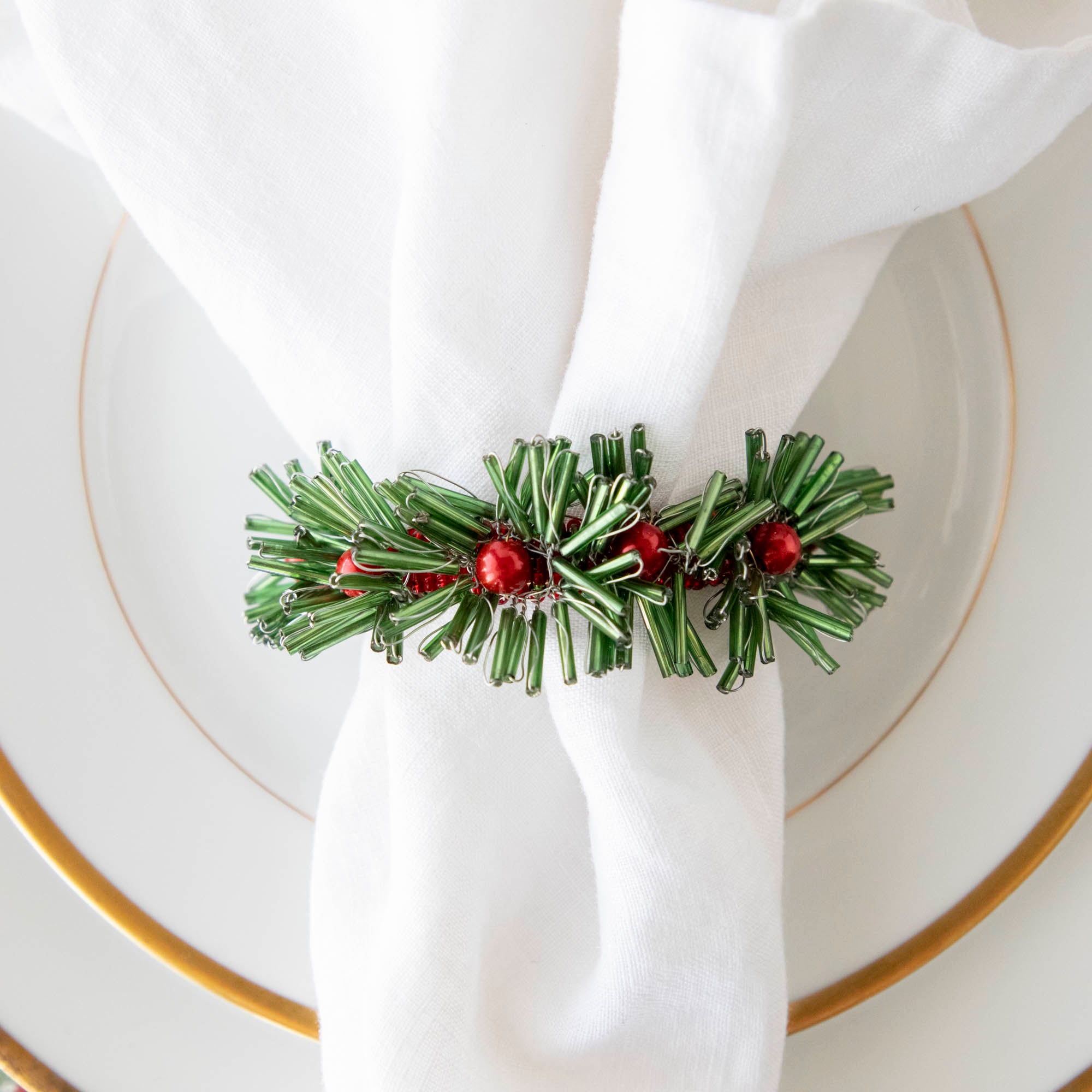 A green and red beaded flower napkin ring on a white napkin, placed on a white plate with gold trim.