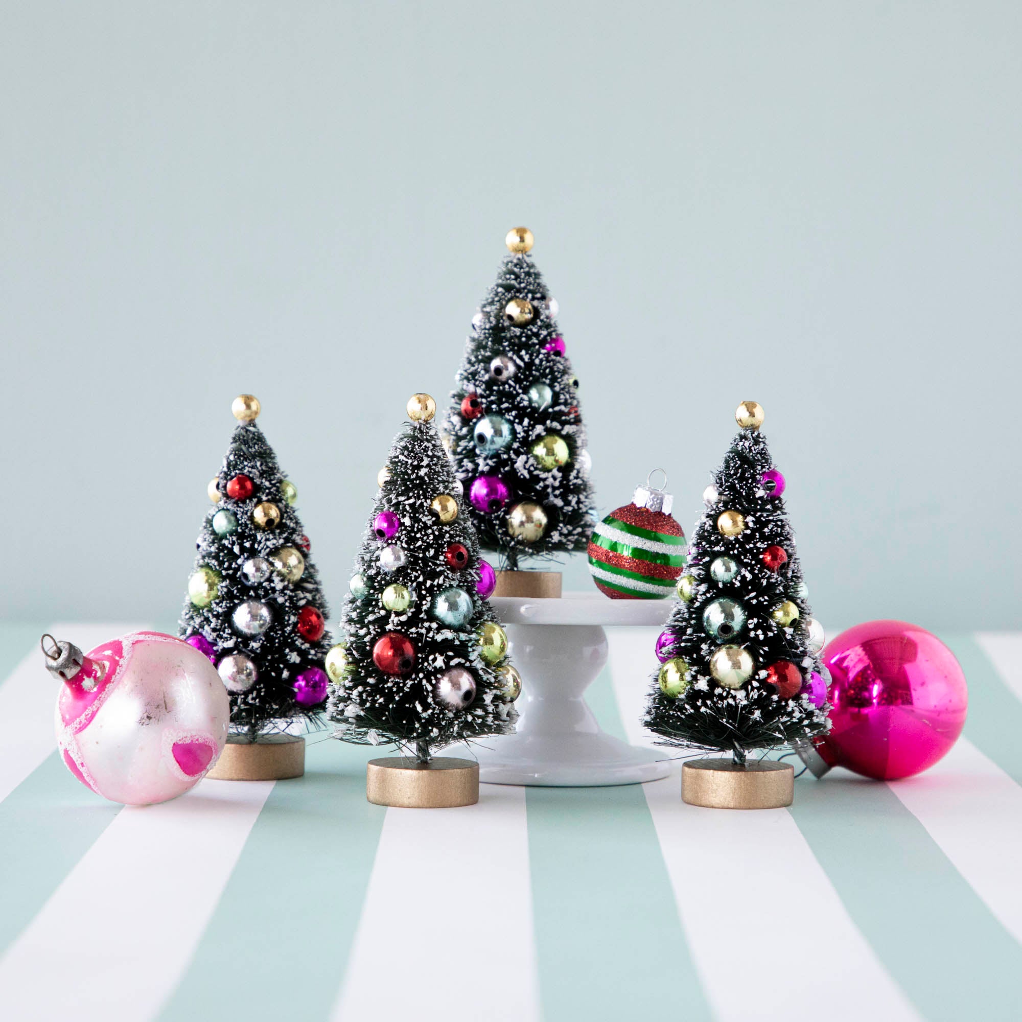Four, 4" holiday sisal trees with colorful ornaments and snow, displayed on a table with ornaments.