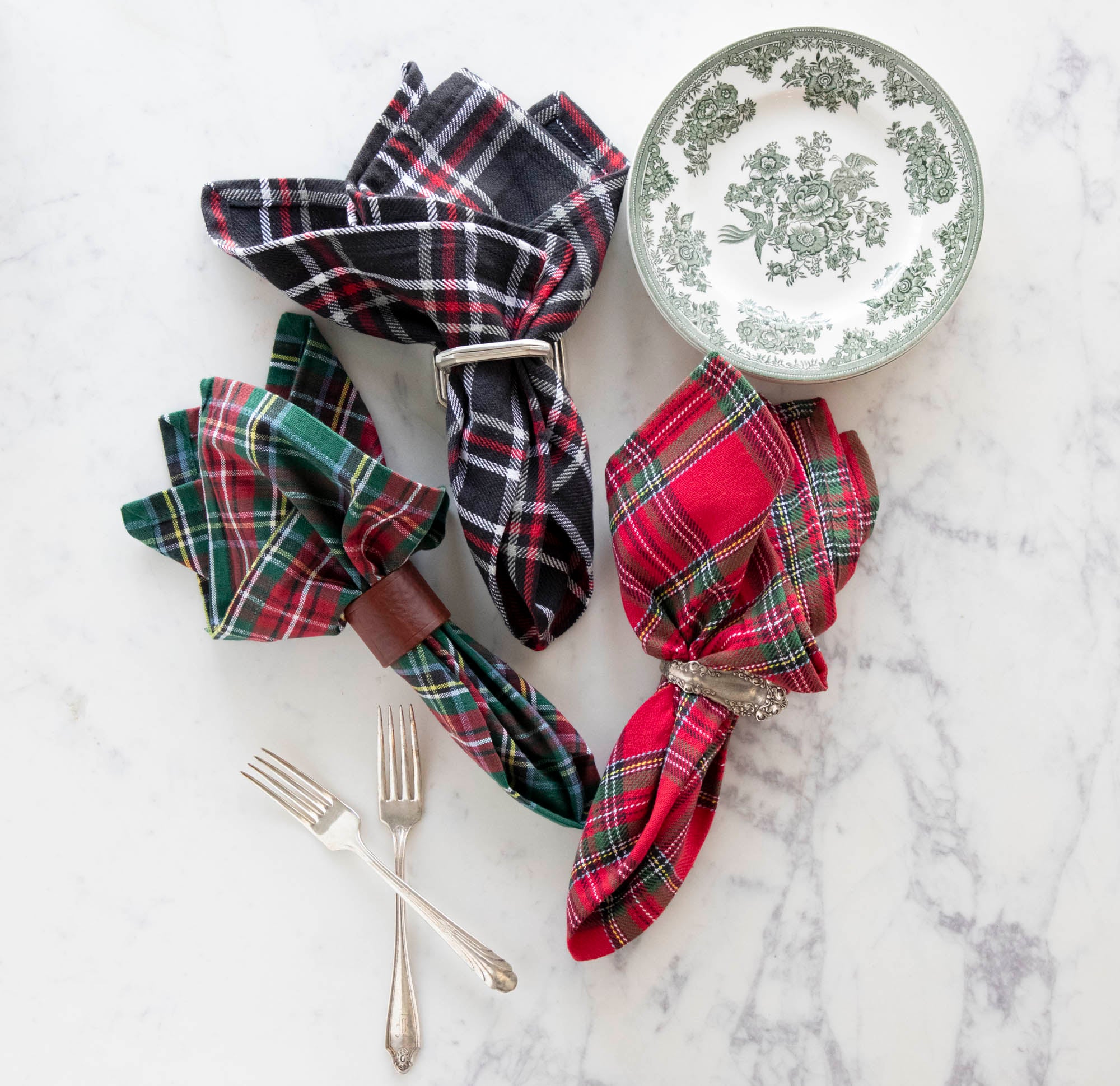 Three different plaid napkins in napkin rings, displayed on a white table with forks and a plate.