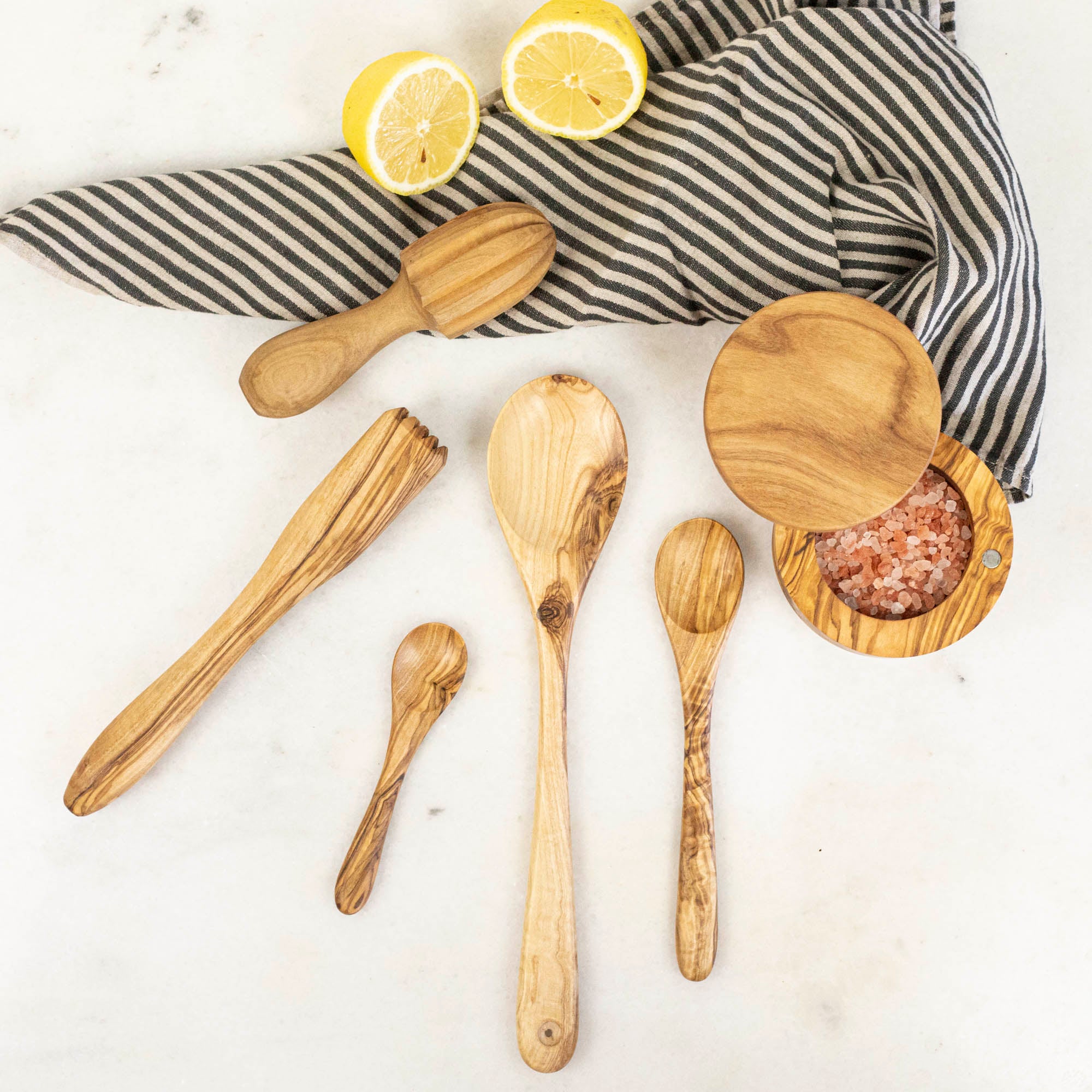 Olive wood kitchen tools, including a juicer, pinch pot, muddler and spoons, on a marble table with a sliced lemon and cloth napkin.