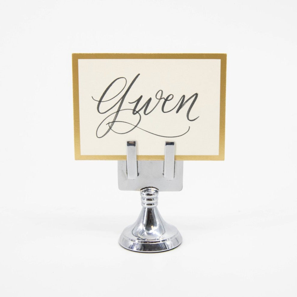 An elegant, chrome, short round stand topped by a flat card-holder with two flat prongs, holding a place card reading &quot;Gwen&quot;.