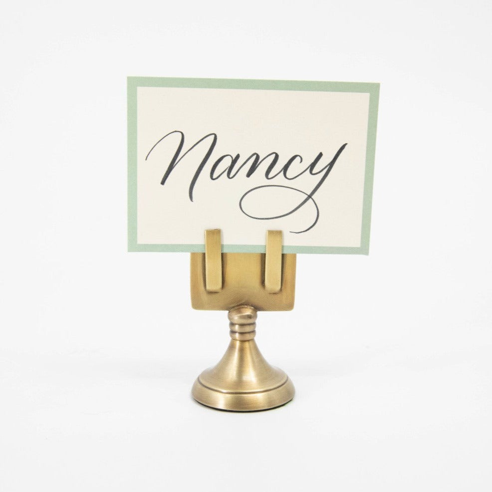 An elegant, brass, short round stand topped by a flat card-holder with two flat prongs, holding a place card reading &quot;Nancy&quot;.