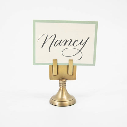 An elegant, brass, short round stand topped by a flat card-holder with two flat prongs, holding a place card reading &quot;Nancy&quot;.