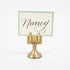 An elegant, brass, short round stand topped by a flat card-holder with two flat prongs, holding a place card reading "Nancy".