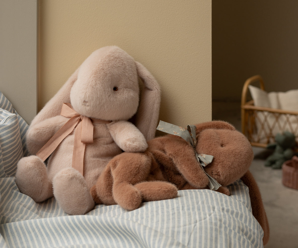 Two Maileg Plush Sleeping Bunnies, one sitting up, one laying down, on a bed in a child&