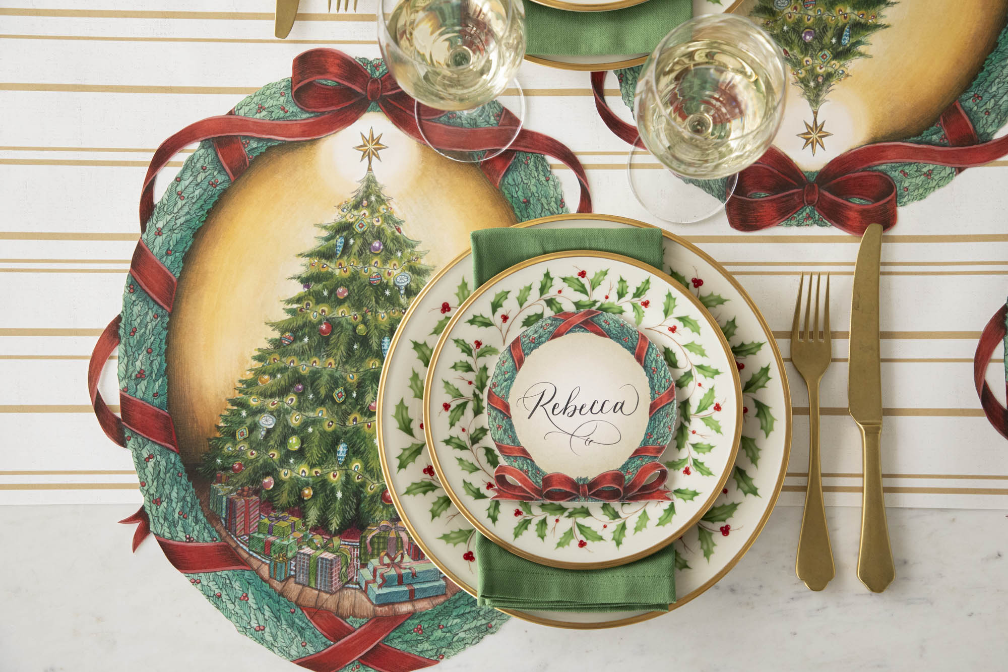 Elegant table setting with the O Christmas Tree Wreath Placemat under Holly dinner plates, gold silverware and a O Christmas Tree Wreath Place Card with the name "Rebecca" written on it.