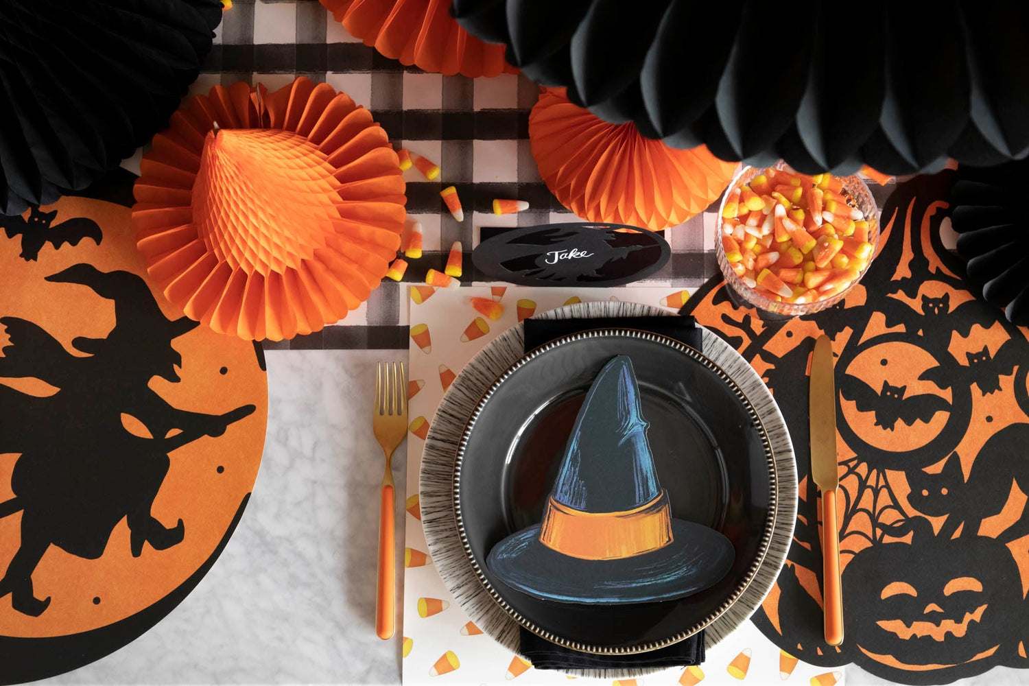 A Halloween-themed table setting from our Halloween Collection includes the Witch Hat Table Accent on a plate, orange and black decorations, a candy corn bowl, and utensils arranged on the Black Painted Check Runner.