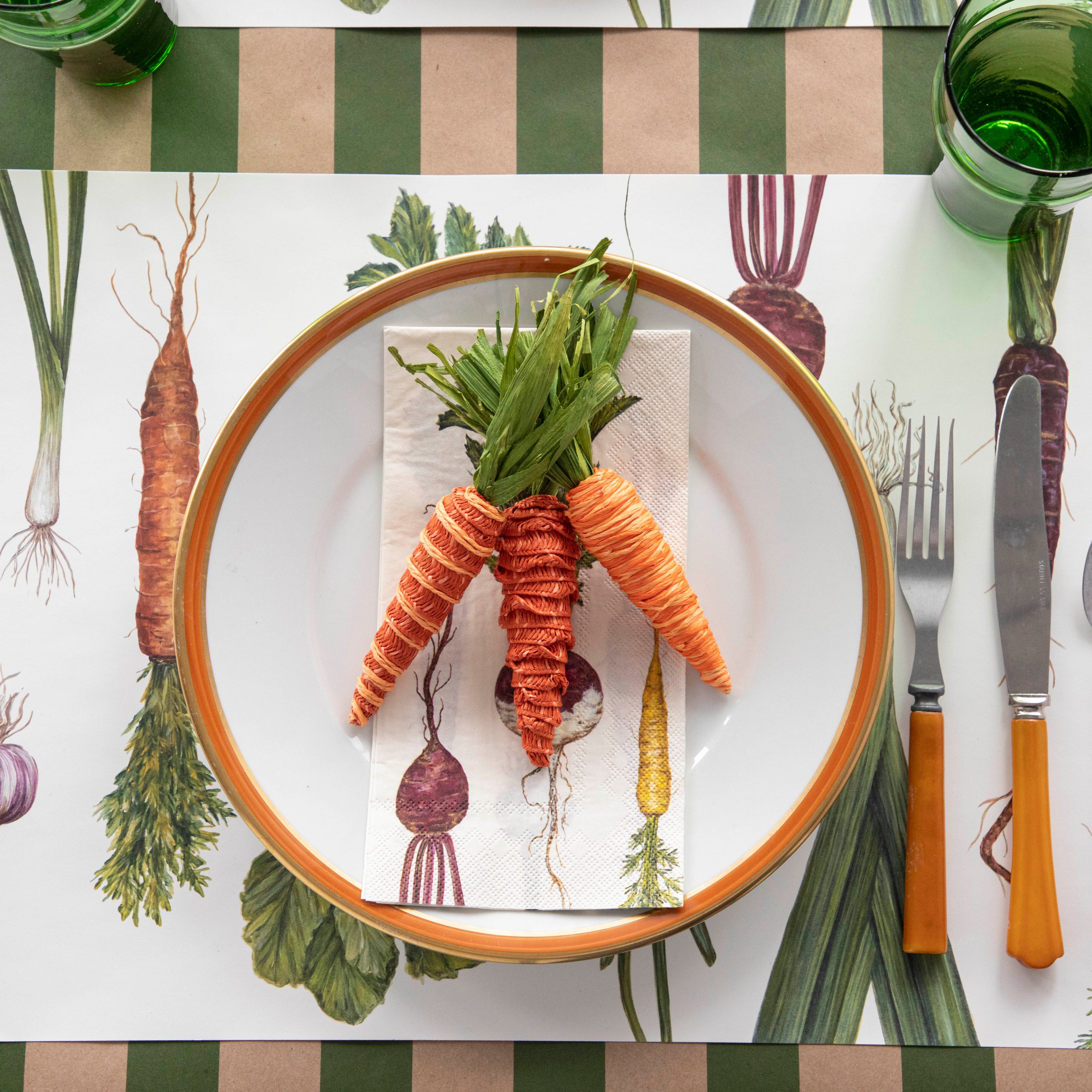 A plate of fresh carrots on a table with a vegetable-themed tablecloth, flanked by cutlery with orange handles.