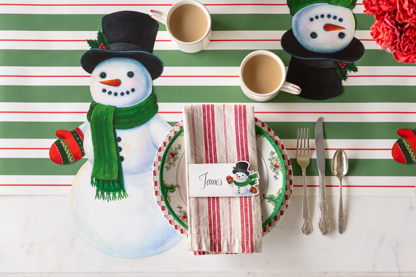 A festive place setting with the Die-cut Snowman Placemat, Snowman Place Card centered on the plates on top of a napkin and Green & Red Awning Stripe Runner underneath.