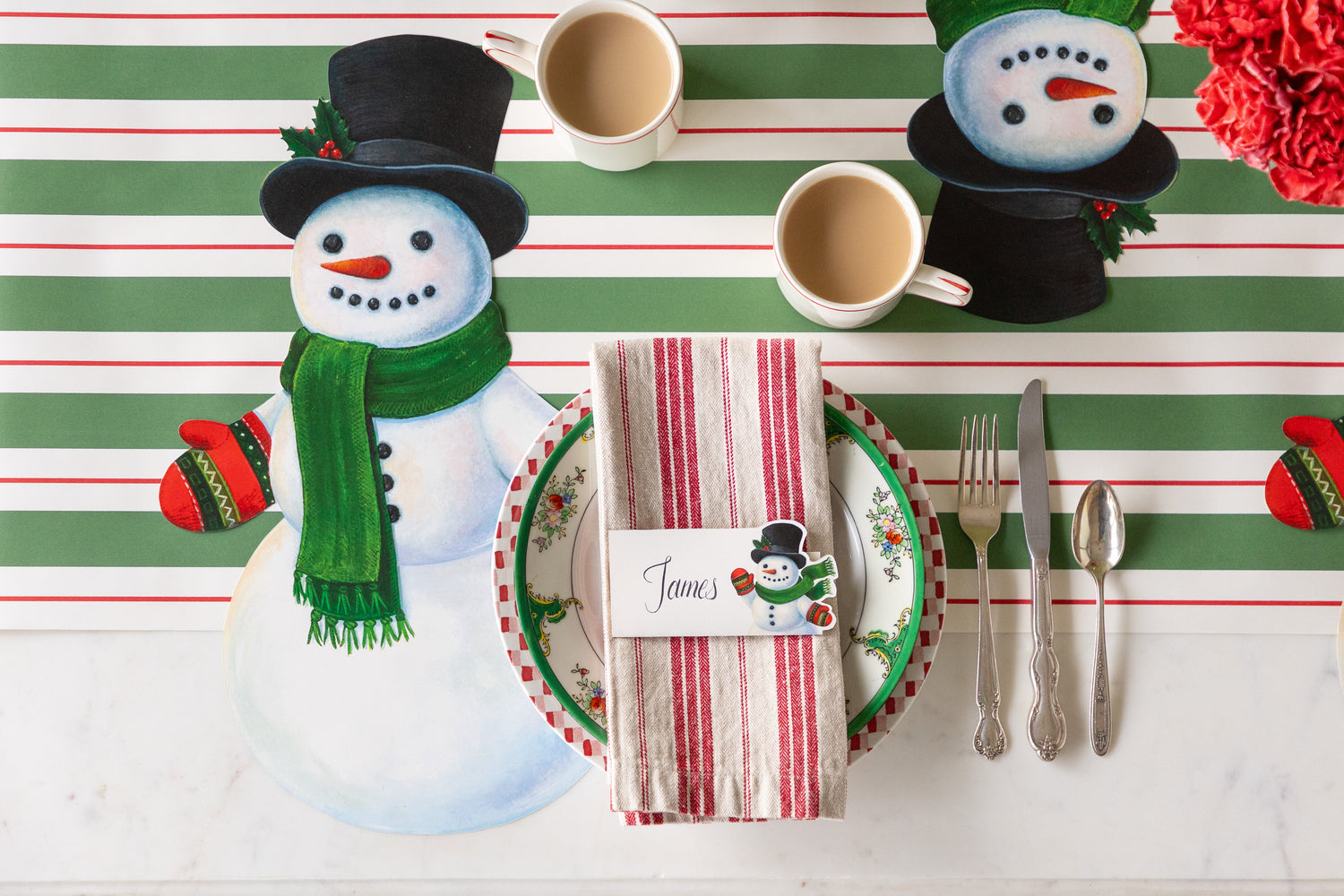 A festive place setting with the Die-cut Snowman Placemat, Snowman Place Card centered on the plates on top of a napkin and Green &amp; Red Awning Stripe Runner underneath.