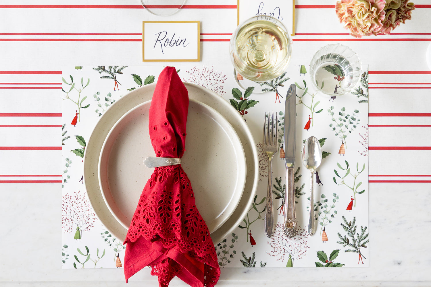 A Christmas themed place setting featuring the Happy Christmas Placemat, vintage silverware, off-white dinnerware, a red lace napkin, and gold frame place card on the Antique Red Stripe Runner.