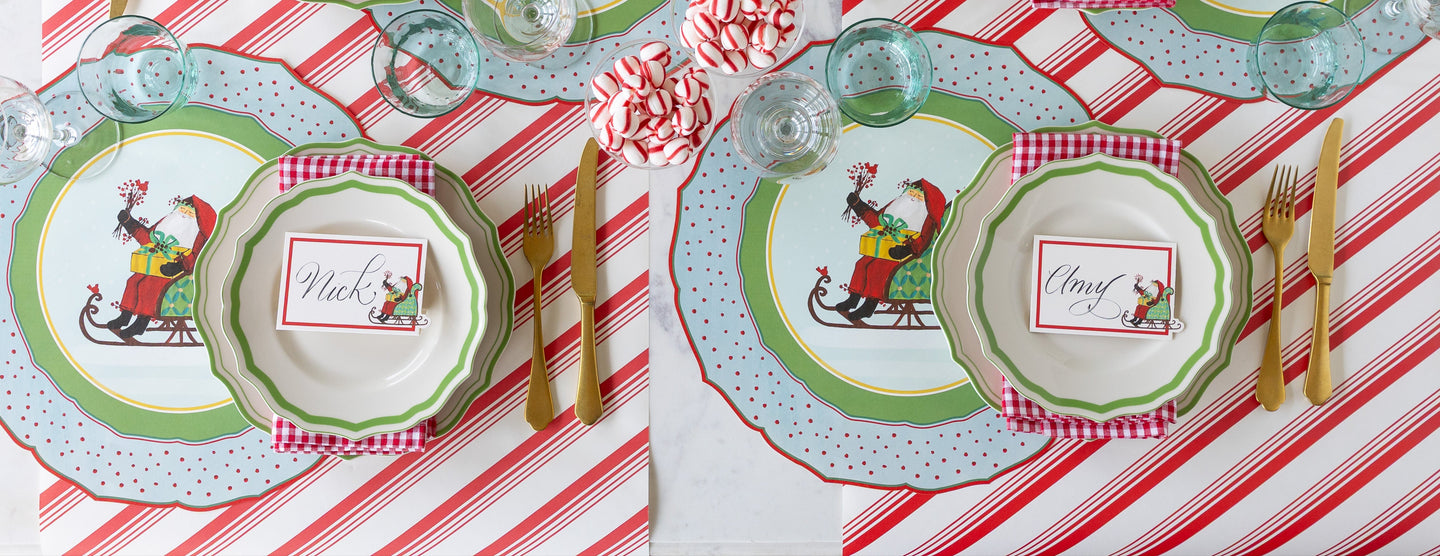 A Christmas themed table setting featuring the Die-cut Old St. Nick Sleigh Placemats, Green Icing on the Cake plates, Old St. Nick Sleigh Place Cards and gold flatware on the Peppermint Stripe Runner.