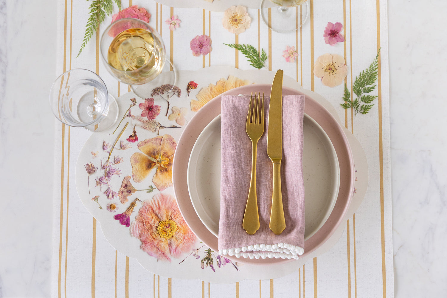 A spring table setting featuring the Die-cut Periwinkle Petals Placemat, floral decor, pink and cream plates and gold flatware on the Antique Gold Stripe Runner.
