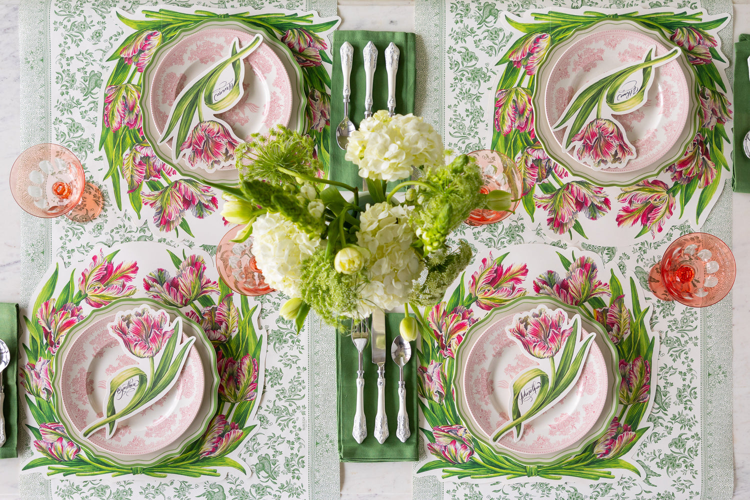 A Spring table setting with a floral centerpiece, Die-cut Parrot Tulip Placemats, Parrot Tulip Table Accents, pink dinner plates and green napkins on the Green Regal Peacock Runner.