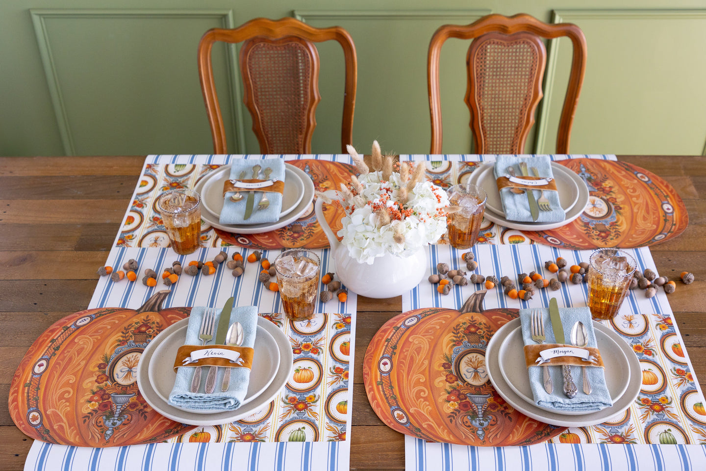 An Autumnal table setting that has 4 placesettings with the Die-Cut Antique Pumpkin Placemat at each along with cutlery, glassware and napkins.  There are acorns down the center of the table and a pitcher with flowers in it.  