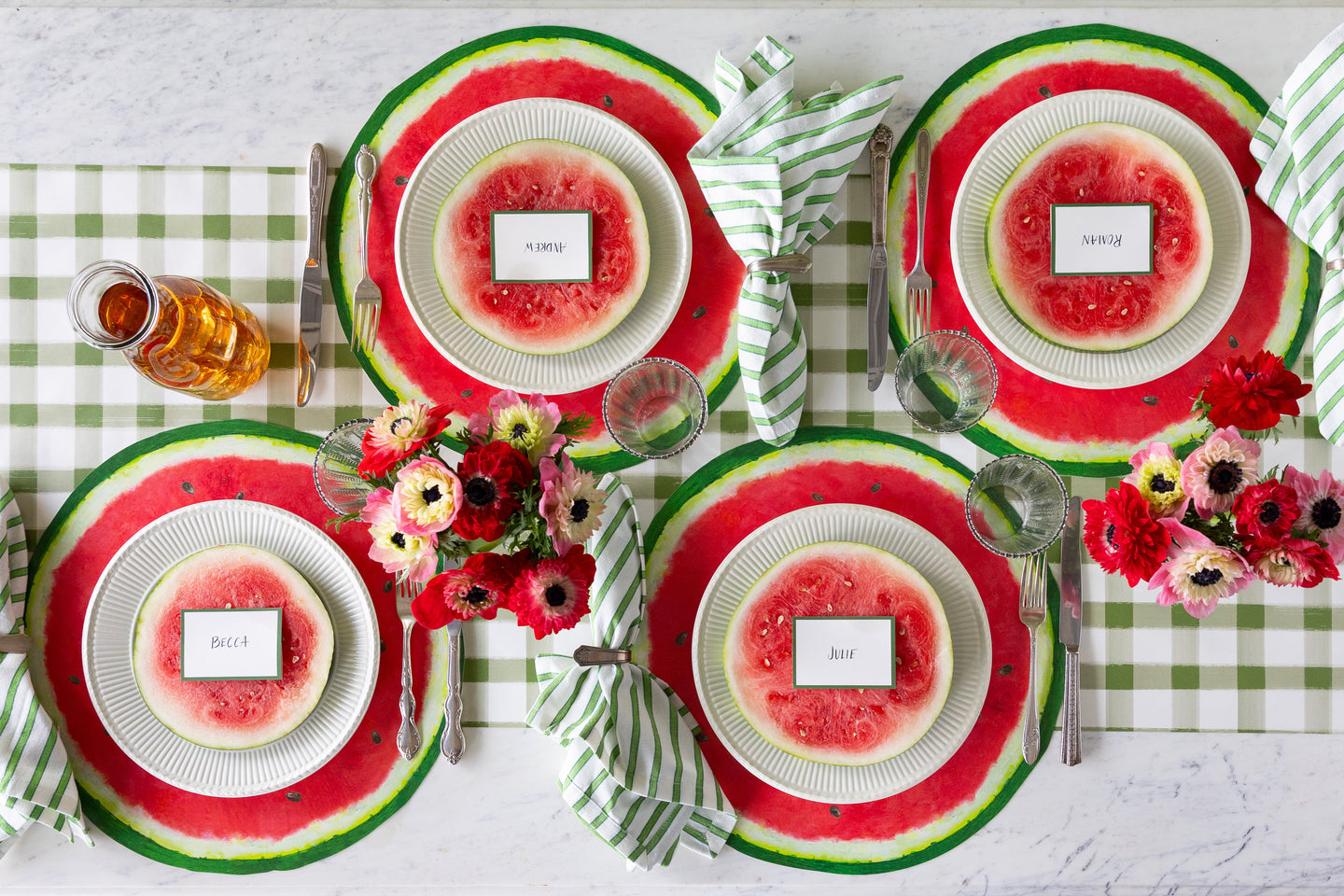 A picnic table setting featuring the Die-cut Watermelon Placemat, flowers, watermelon slices and green striped napkins on the Moss Painted Check Runner.
