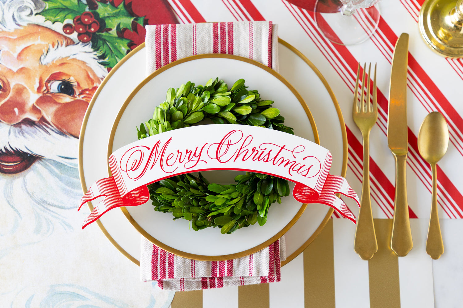 A Christmas themed place setting featuring the Red Merry Christmas Banner Table Accent atop a topiary wreath, gold rimmed plates and a red-and-white striped napkin.