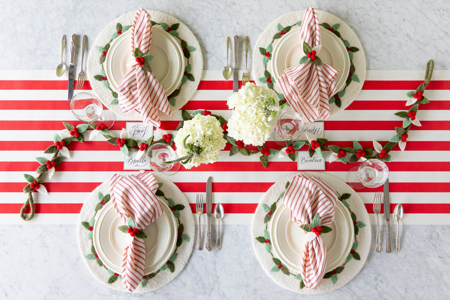 A festive table setting featuring the Felt Berry Wreath Placemats, Felt Holly Napkin Rings, red-and-white striped napkins, flowers and Felt Berry Garland on the Red Classic Stripe Runner.
