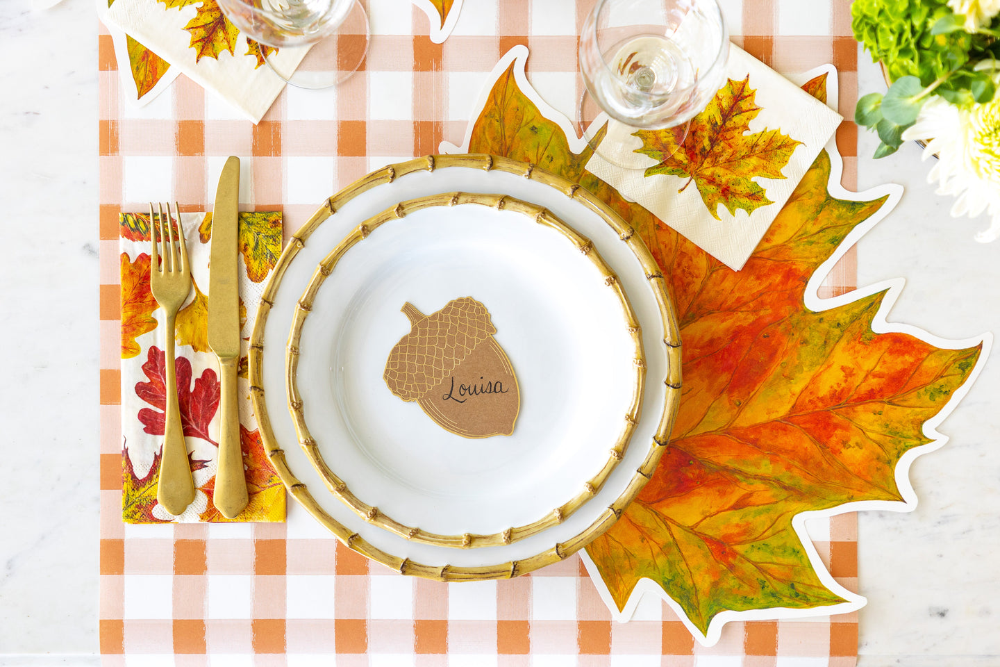 An elegant place setting featuring the Die-cut Fall Leaf Placemat, Acorn Place Card centered on dinner plates, Fall Foliage Cocktail and Guest Napkins and Orange Painted Check Runner underneath.