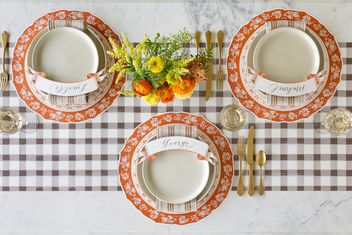 A fall table setting for three with a floral centerpiece, featuring the Die-cut Pumpkin Regal Placemat, gold flatware, wine glasses, salt dinnerware and Orange Banner Table Accent on the Brown Painted Check Runner.