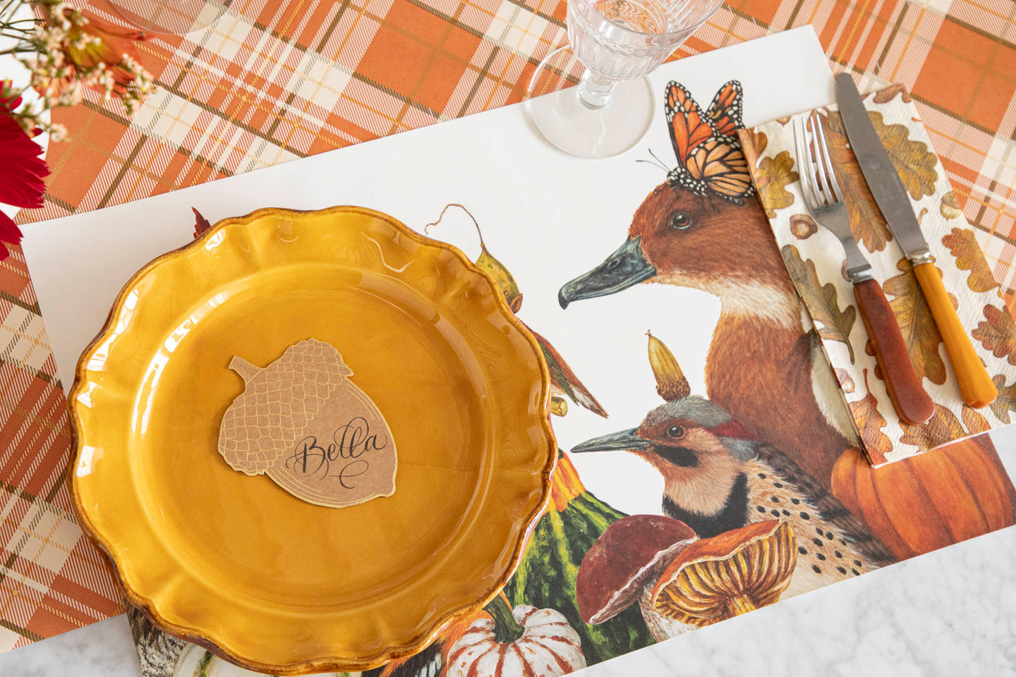 A fall place setting featuring the We Gather Together Placemat with the Autumn Leaves Guest Napkin, vintage flatware and dinner plate and Acorn Place Card atop, on the Orange Plaid Runner.