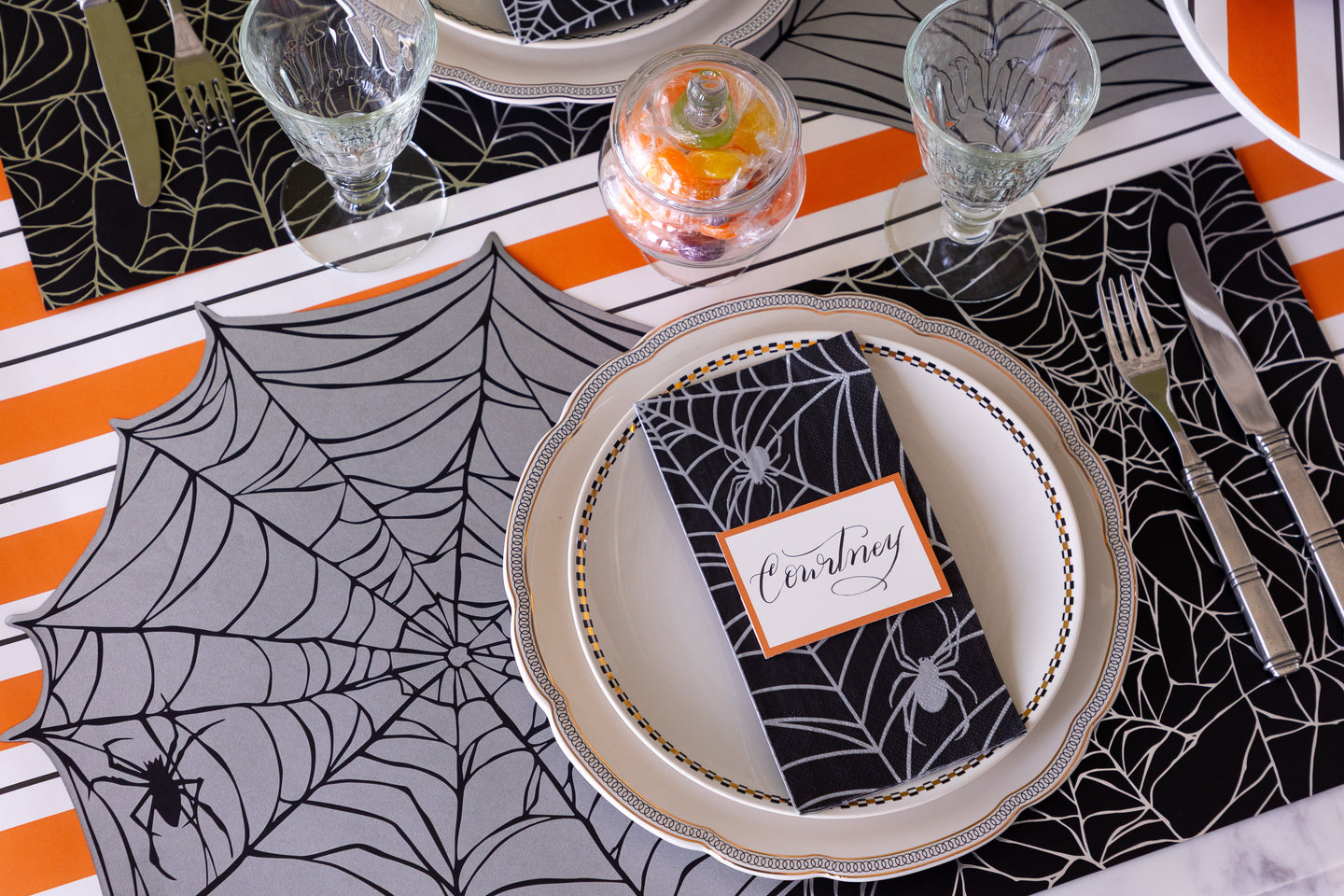 A Halloween themed table setting featuring the Die-cut Spiderweb Placemat, Spiderweb Placemat, Spiderweb Guest Napkin and Orange & Black Awning Stripe Runner underneath.