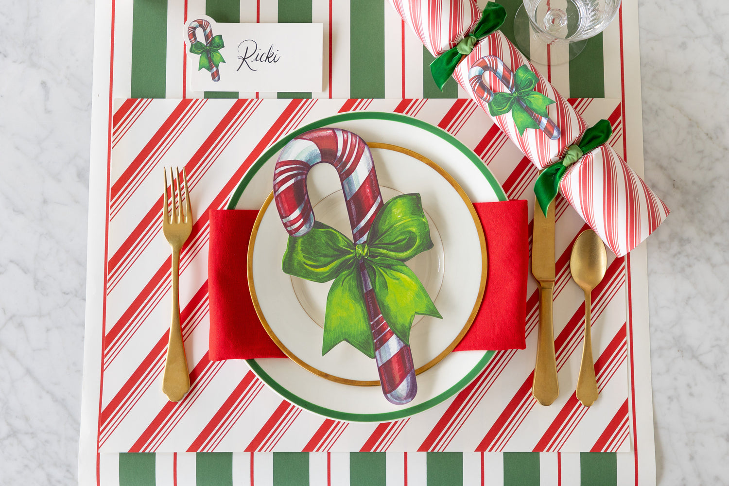 A Christmas place setting featuring the Peppermint Stripe Placemat, a Candy Cane Cracker, Candy Cane Table Accent atop plates, and Candy Cane Place Card on the Green &amp; Red Awning Stripe Runner.