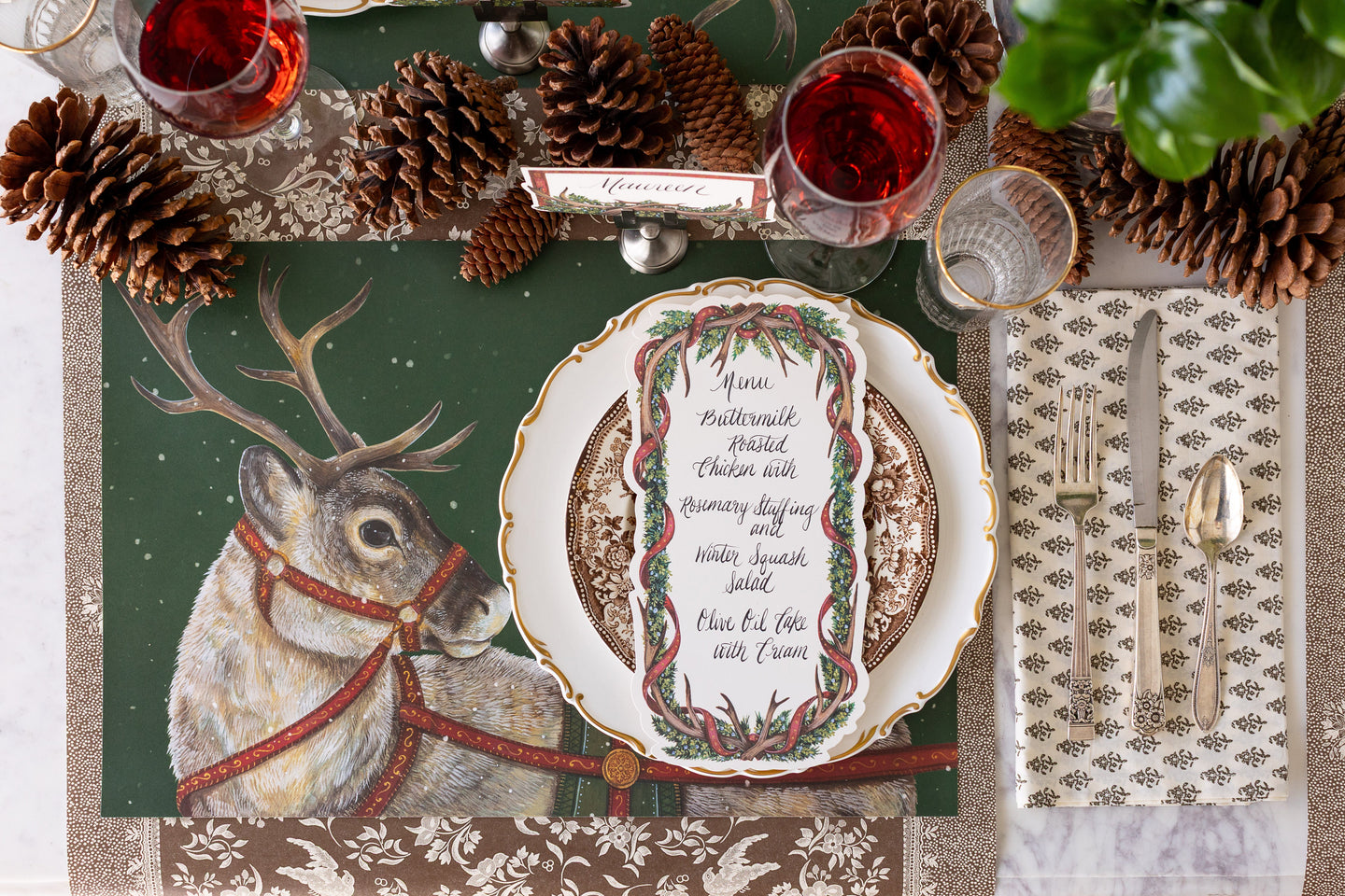 A woodland table setting featuring the Holiday Reindeer Placemat, Antler Garland Table Accent, a floral napkin and vintage silverware on the Brown Regal Peacock Runner.